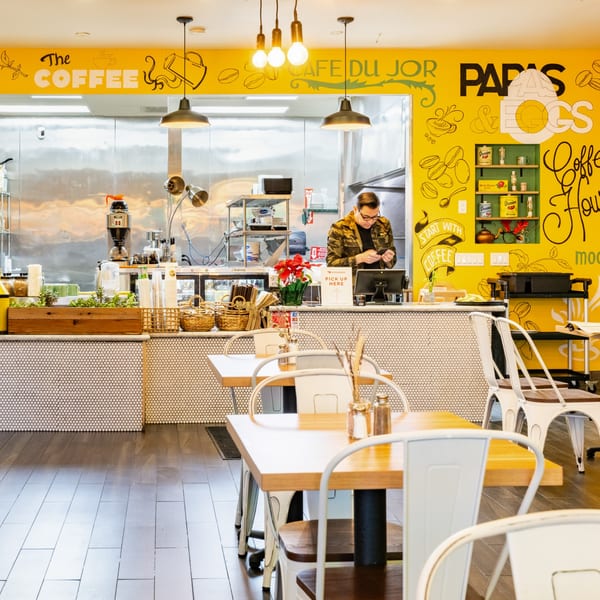 A bright yellow breakfast restaurant interior with a counter, a person working, and dining tables.