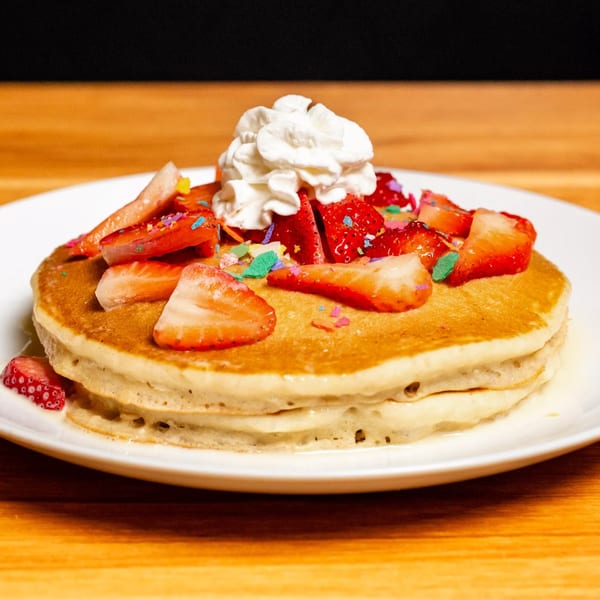 Fluffy pancakes topped with fresh strawberries, whipped cream, and colorful sprinkles on a white plate.