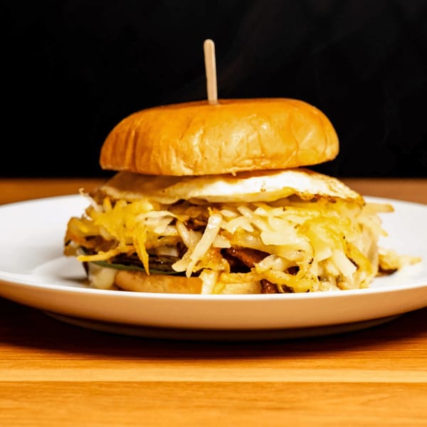 A breakfast sandwich with a fried egg, hash browns, and meat on a bun, served on a white plate.