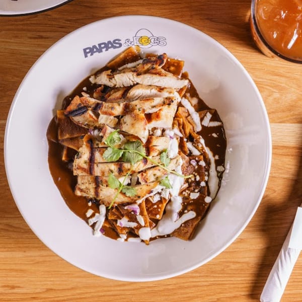 Papas and Eggs chilaquiles with grilled chicken, dark sauce, crema, and cilantro on a white plate.