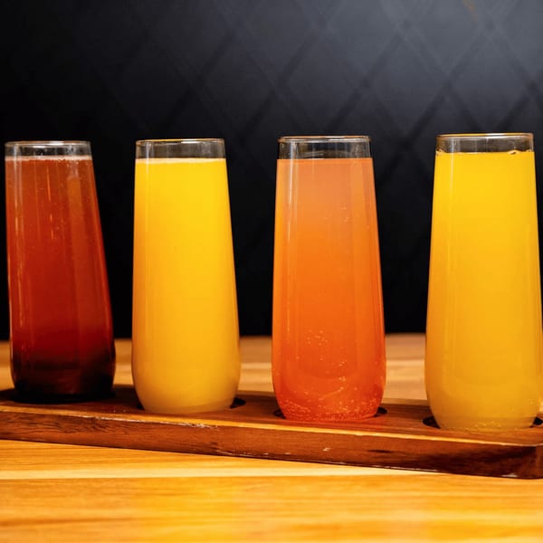 Four colorful mimosa flights, including orange, grapefruit, and cranberry, on a wooden tray.