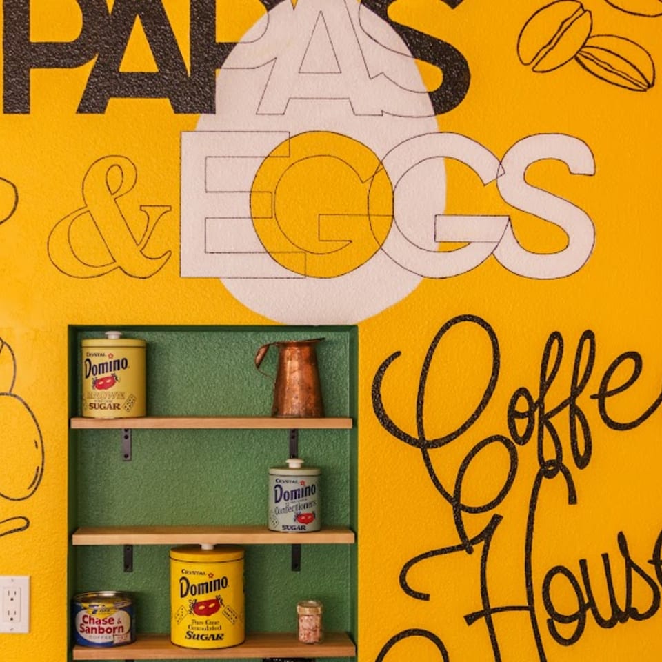 A yellow wall with "Papas & Eggs Coffee House" text and shelves holding vintage sugar and coffee cans.