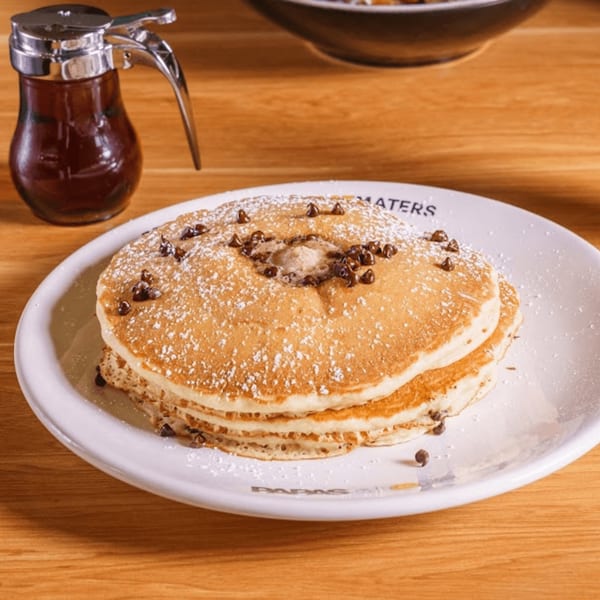Stack of Papas and Eggs chocolate chip pancakes with powdered sugar, butter, and a syrup dispenser.